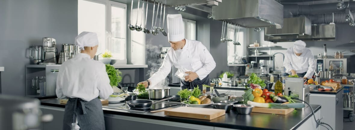 widok na kuchnię, trzech kucharzy pracuje pośród palników, przyborów kuchennych i warzyw